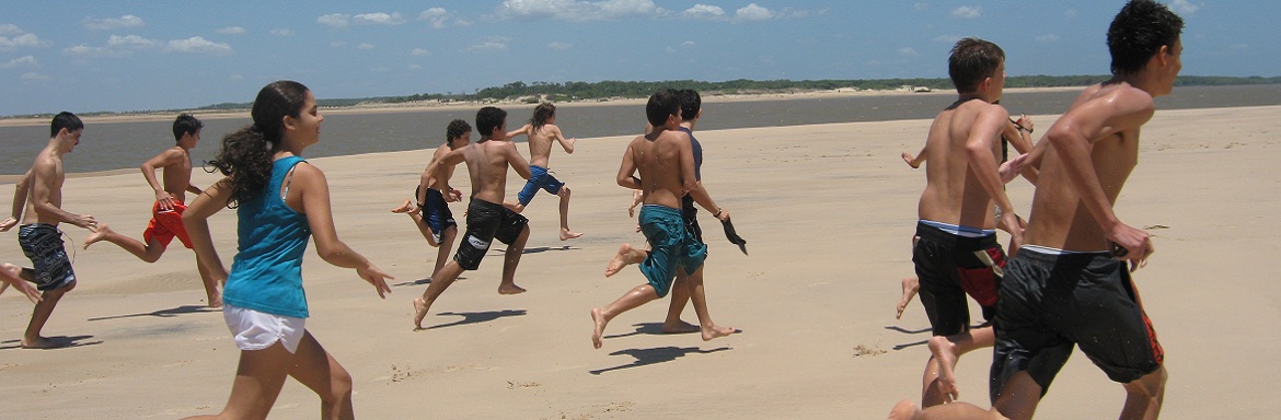 Na imagem, crianças correm pela areia da praia. O céu está azul e nuvens.