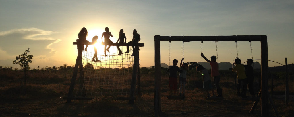 A fotografia mostra crianças brincando em um balanço e em um trepa-trepa. Elas estão contra a luz durante o pôr-do-Sol