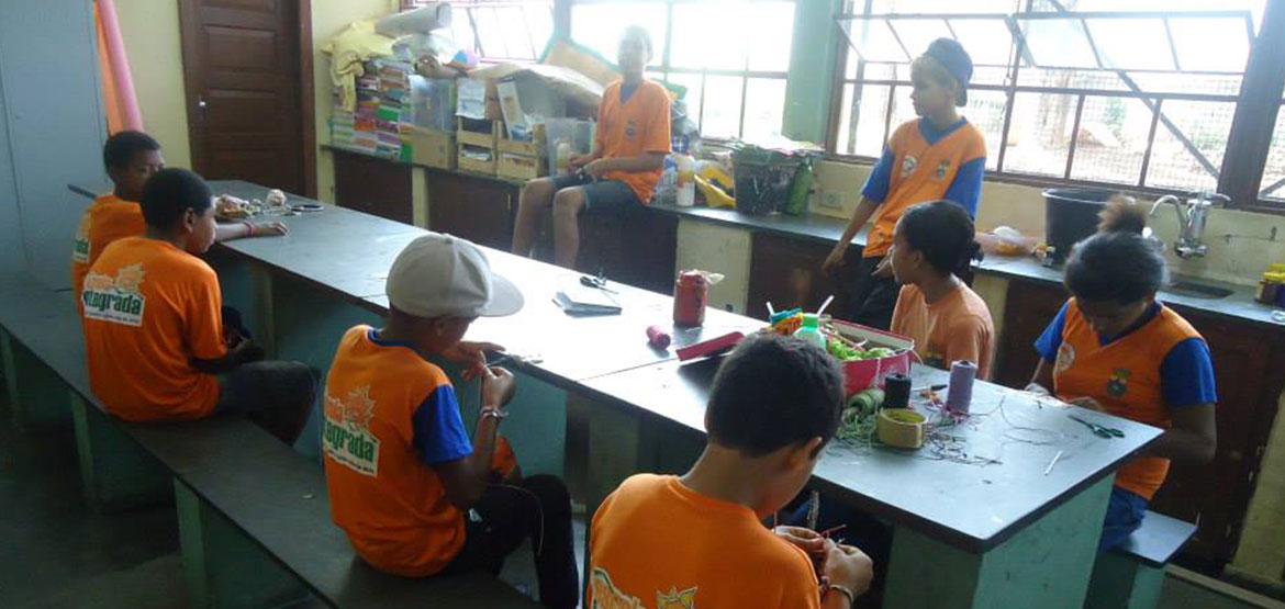 Um grupo de crianças está sentada em uma grande mesa retangular. Elas estão utilizando materiais de pintura.
