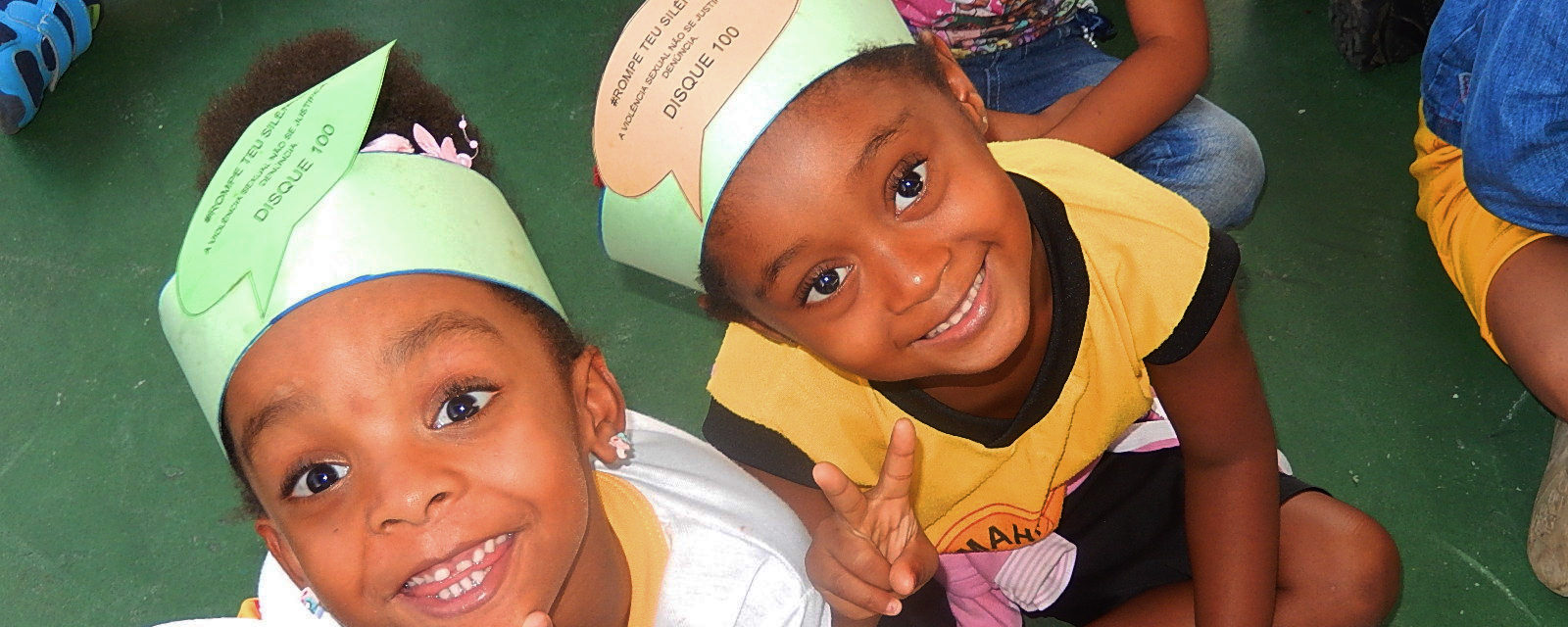 Na foto estão duas crianças sorrindo. A câmera está posicionada no alto. As crianças usam uma faixa de papel em volta da cabeça.