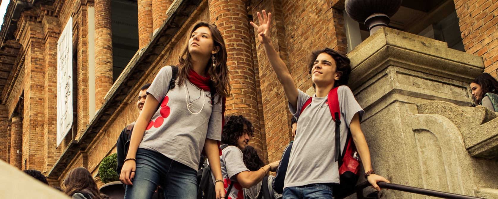 Dois estudantes observam o horizonte. Um deles acena. Ao fundo, um casarão de tijolos.