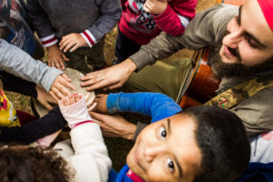 a imagem mostra um grupo de crianças e um adulto. Eles estão em roda e com um dos braços esticados para frente, tocando um objeto comum.